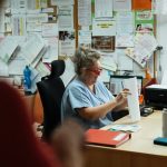 A senior caregiver in blue scrubs sorting paperwork at a hospital office desk.